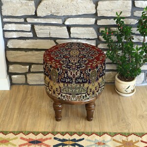 May include: A circular, upholstered footstool with a detailed, patterned top in red, blue, and gold. The sides feature an elephant motif. The footstool stands on four short, wooden legs. A small potted plant is nearby.