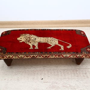 May include: A rectangular, red footstool with a detailed lion design in the center. The lion is beige and the stool has a decorative border. The stool is supported by four short, dark wooden legs. A small potted plant is to the right.
