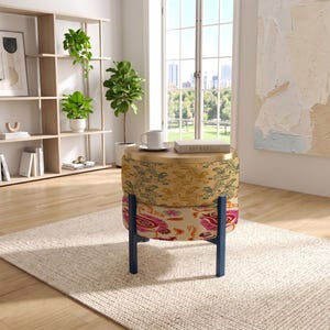 May include: A round ottoman with a flat wooden top, featuring a two-tiered upholstered base with a yellow floral pattern on the upper section and a pink and red paisley print on the lower section, supported by four dark blue wooden legs.