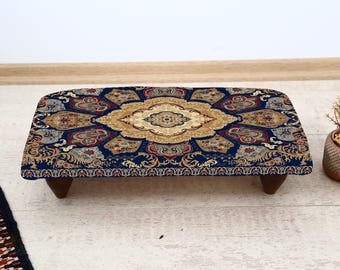 May include: A rectangular footstool with a patterned top in navy blue, gold, and red. The design features an ornate, symmetrical pattern. The footstool has four short, dark brown wooden legs.