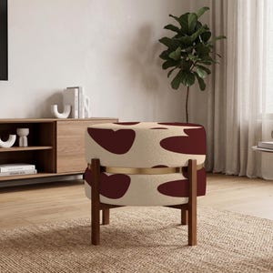 May include: A round ottoman featuring a cream-colored fabric with a dark red abstract cow-print pattern. The seat is encircled by a thin gold-toned metal band and is supported by four straight, dark brown wooden legs in a simple, sturdy design.
