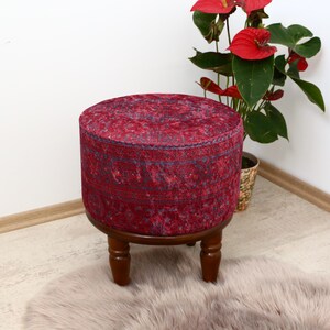 May include: A red and blue patterned ottoman with a wooden base and three legs. The ottoman is sitting on a fluffy white rug.