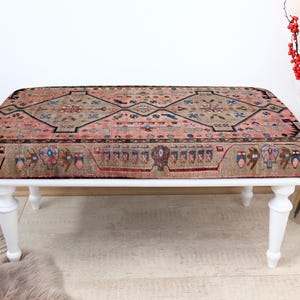 May include: A white wooden bench with a pink and beige patterned rug top. The rug has a geometric design with intricate details.