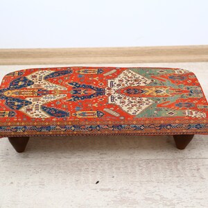 May include: A rectangular ottoman with a vibrant, patterned textile top in shades of red, blue, and cream. The ottoman rests on four dark wooden legs. The design is reminiscent of a traditional rug.
