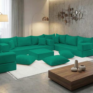 May include: A large, modular emerald green sectional sofa with multiple cushions and ottomans. The sofa is arranged in an L-shape, with a matching square ottoman and several throw pillows. A wooden coffee table and decorative vases are in front.