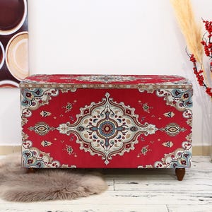 May include: A red storage ottoman with a colourful patterned fabric design. The ottoman has a rectangular shape and wooden legs.