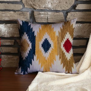 May include: Decorative throw pillow with a geometric pattern in navy blue, yellow, and red diamonds on a white background. The pillow is square and rests on a wooden surface, with a stone wall backdrop.