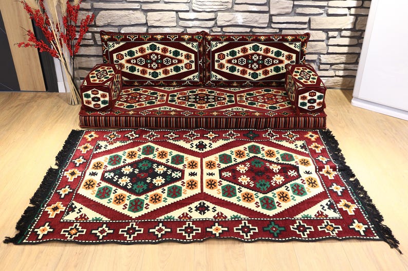 Op de afbeelding: Een patroon vloerkleed in rood, cr&egrave;me en zwart, met een bijpassende bank en kussens. De bank heeft een geometrisch ontwerp met rode, groene en gouden accenten. Het vloerkleed heeft een zwarte franje.