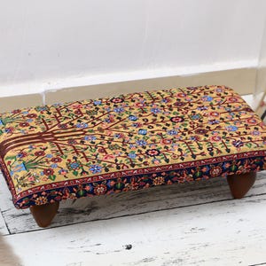 May include: A rectangular footstool with a colorful floral pattern on a tan background. The design includes a tree motif with blue, red, and green accents. The stool has dark brown wooden legs and a dark blue border.