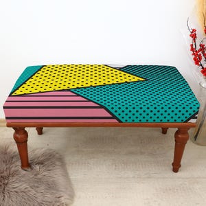 May include: A small footstool with a vibrant, geometric design. The top is patterned with yellow, teal, and pink shapes, outlined in black. The wooden legs are a dark brown colour. The stool is approximately 60 cm long.