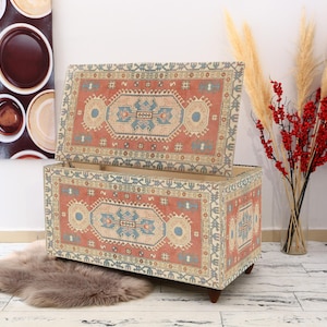 May include: A decorative storage ottoman with a colorful patterned fabric cover. The ottoman is open, revealing a wooden interior. The fabric features a traditional rug design with red, blue, and cream colors.