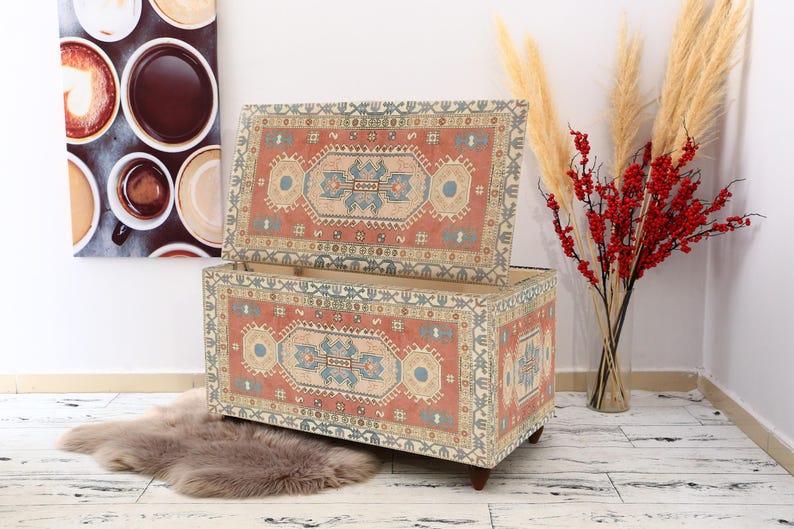 May include: A decorative storage ottoman with a colorful patterned fabric cover. The ottoman is open, revealing a wooden interior. The fabric features a traditional rug design with red, blue, and cream colors.