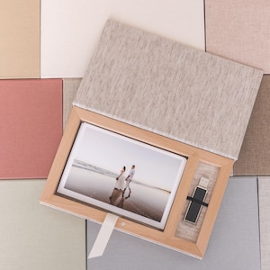Puede incluir: Una caja de madera con una tapa de lino, que contiene una fotografía enmarcada de una pareja caminando por la playa y una unidad USB. La caja está rodeada de muestras de tela de varios colores.