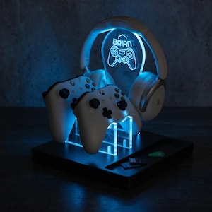 May include: A black gaming station with blue LED lighting, holding two white game controllers and a pair of white headphones. The station has a clear acrylic panel with the name "BRIAN" and a game controller symbol. A small tray holds accessories.