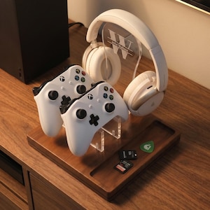 May include: A wooden gaming accessory organizer with two white game controllers, a white headset, and a clear acrylic stand. The organizer also holds a guitar pick and memory cards. The background includes a black game console.