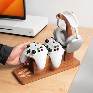May include: A wooden gaming accessory stand holds two white game controllers and a pair of white headphones. The stand is brown with a monogrammed design. The setup is on a light wood surface.