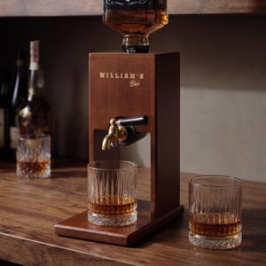 Puede incluir: Un dispensador de licor de madera con un grifo de lat&oacute;n y el texto "WILLIAM'S Bar". Dos vasos de cristal con l&iacute;quido &aacute;mbar sobre una superficie de madera. Una botella de licor se coloca encima.