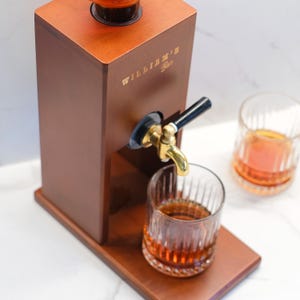 Puede incluir: Un dispensador de licores de madera con un grifo de lat&oacute;n, que dispensa l&iacute;quido &aacute;mbar en un vaso. El dispensador tiene el texto "WILLIAM'S Bar" en letras doradas. Se ven dos vasos de l&iacute;quido &aacute;mbar.