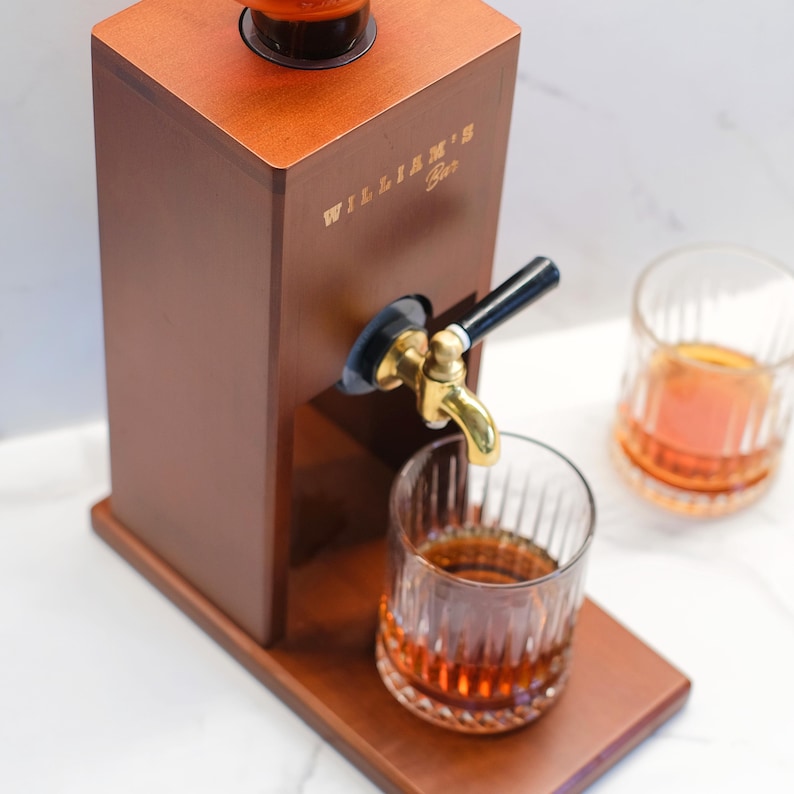 Puede incluir: Un dispensador de licores de madera con un grifo de lat&oacute;n, que dispensa l&iacute;quido &aacute;mbar en un vaso. El dispensador tiene el texto "WILLIAM'S Bar" en letras doradas. Se ven dos vasos de l&iacute;quido &aacute;mbar.