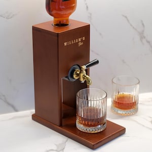 Puede incluir: Un dispensador de licores de madera con un grifo de lat&oacute;n y el texto "WILLIAM'S Bar". El dispensador contiene una botella de l&iacute;quido &aacute;mbar y se encuentra sobre una base de madera. Dos vasos de l&iacute;quido &aacute;mbar est&aacute;n junto al dispensador.