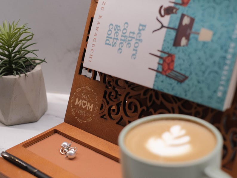 May include: A wooden book stand with a laser-cut design. The stand has a small compartment for jewelry or other small items. The stand is engraved with the words "Best Mom Ever". A book titled "Before the Coffee Gets Cold" by  Zukiawaguci is displayed on the stand. A cup of coffee with latte art is in the foreground.
