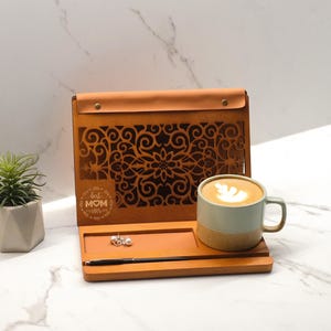 May include: A wooden tray with a cutout design and the words "Best Mom Ever" engraved on the front. The tray has a small compartment for jewelry and a pen holder. A cup of coffee with latte art sits on the tray.
