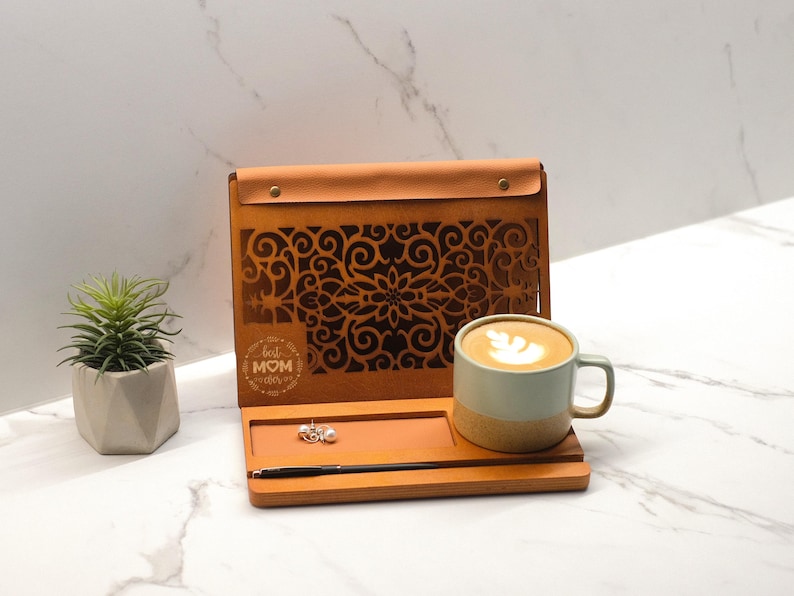 May include: A wooden tray with a cutout design and the words "Best Mom Ever" engraved on the front. The tray has a small compartment for jewelry and a pen holder. A cup of coffee with latte art sits on the tray.