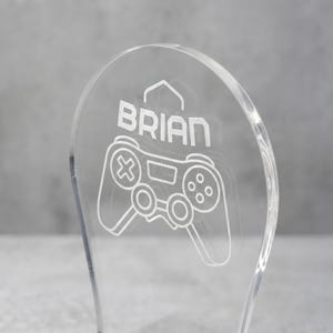 Może przedstawiać: Przezroczysta dekoracja do gier z akrylu z napisem "BRIAN" i białym wzorem kontrolera. Kontroler ma przyciski i joysticki, idealne do pokoju gracza lub na biurko.