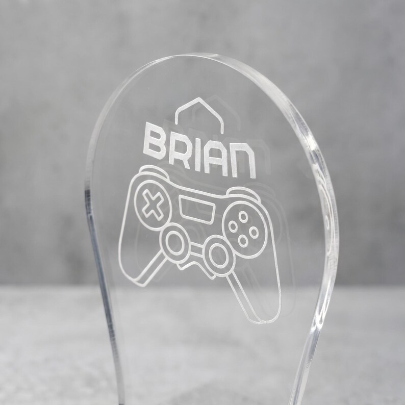 Może przedstawiać: Przezroczysta dekoracja do gier z akrylu z napisem "BRIAN" i białym wzorem kontrolera. Kontroler ma przyciski i joysticki, idealne do pokoju gracza lub na biurko.