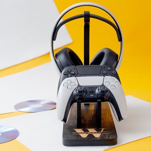 May include: A white and black gaming headset and controller are displayed on a wooden stand. The stand has the word "Gaming" engraved on it. The headset is resting on the stand with the controller in front of it.