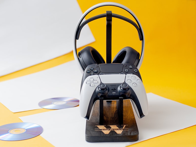May include: A white and black gaming headset and controller are displayed on a wooden stand. The stand has the word "Gaming" engraved on it. The headset is resting on the stand with the controller in front of it.