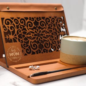 May include: A wooden tray with a leather flap, featuring a laser-cut design of swirling vines and the text "Best Mom Ever". The tray has a built-in stand and includes a small cup of coffee and a pair of earrings.