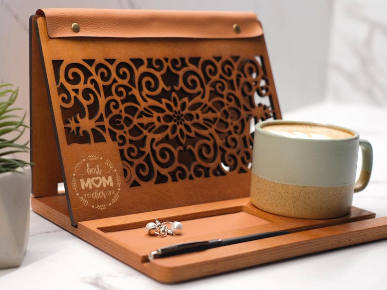 May include: A wooden tray with a leather flap, featuring a laser-cut design of swirling vines and the text "Best Mom Ever". The tray has a built-in stand and includes a small cup of coffee and a pair of earrings.