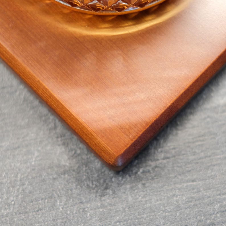 Puede incluir: Una bandeja cuadrada de madera con un acabado marr&oacute;n rojizo c&aacute;lido. La bandeja tiene una superficie lisa y bordes redondeados. Un vaso con un dise&ntilde;o estampado es parcialmente visible en la bandeja. La bandeja est&aacute; sobre un fondo gris.