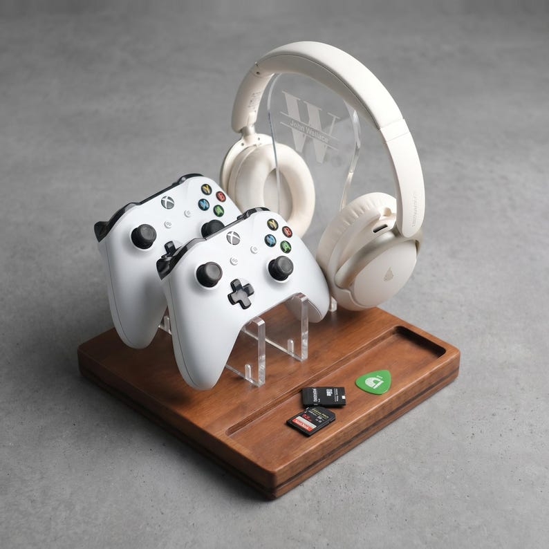 Może przedstawiać: Drewniana stacja do gier z dwoma białymi kontrolerami Xbox, parą białych słuchawek i przezroczystym akrylowym stojakiem. Stacja zawiera r&oacute;wnież kostkę do gitary i dwie karty pamięci. Słuchawki spoczywają na przezroczystym akrylowym stojaku z inicjałami "JW".