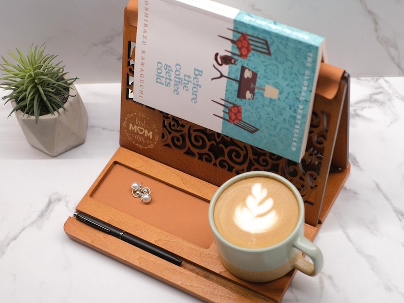 May include: Wooden book stand with a decorative cutout design. The stand has a built-in pen holder, a small tray, and a book holder. The stand is decorated with the words "Best Mom Ever". A book titled "Before the coffee gets cold" is in the book holder. A cup of coffee with latte art is on the tray.