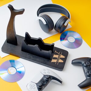 May include: A black wooden stand with a personalised inscription "WW John Wallace" for holding gaming accessories. The stand has two slots for controllers and a hook for headphones. There are two game discs, a black controller, and a white controller on a white surface.