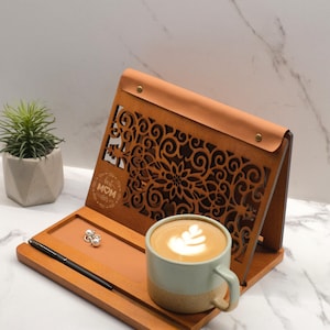 May include: A wooden stand with a decorative cutout design and the text "Best Mom Ever". The stand has a leather-like top and a tray for holding items. A cup of coffee with latte art is on the tray.