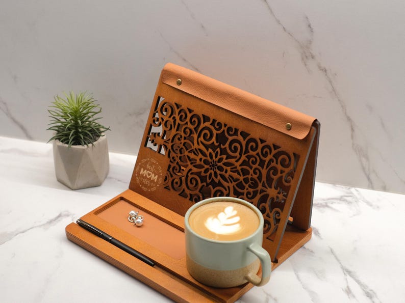 May include: A wooden stand with a decorative cutout design and the text "Best Mom Ever". The stand has a leather-like top and a tray for holding items. A cup of coffee with latte art is on the tray.
