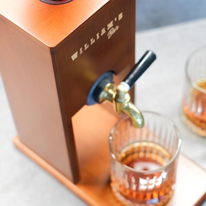 Puede incluir: Un dispensador de licores de madera con un grifo de lat&oacute;n y un mango negro. El dispensador tiene el texto "WILLIAM'S Bar" en el lateral. Un vaso de l&iacute;quido &aacute;mbar se encuentra debajo del grifo, con otro vaso visible en el fondo.