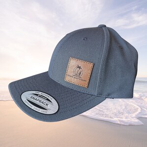 May include: A grey snapback cap with a curved peak. A brown leather patch displays a palm tree and surfboard logo with the text "DOWN SOUTH SURF". The cap has a "Yupoong Since 1974" label.