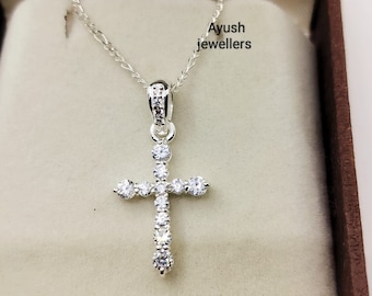 Puede incluir: Un collar de plata con un pequeño colgante de cruz engastado con diamantes. El collar se muestra en una caja de terciopelo marrón con el texto "Ayush jewellers" impreso en el interior.