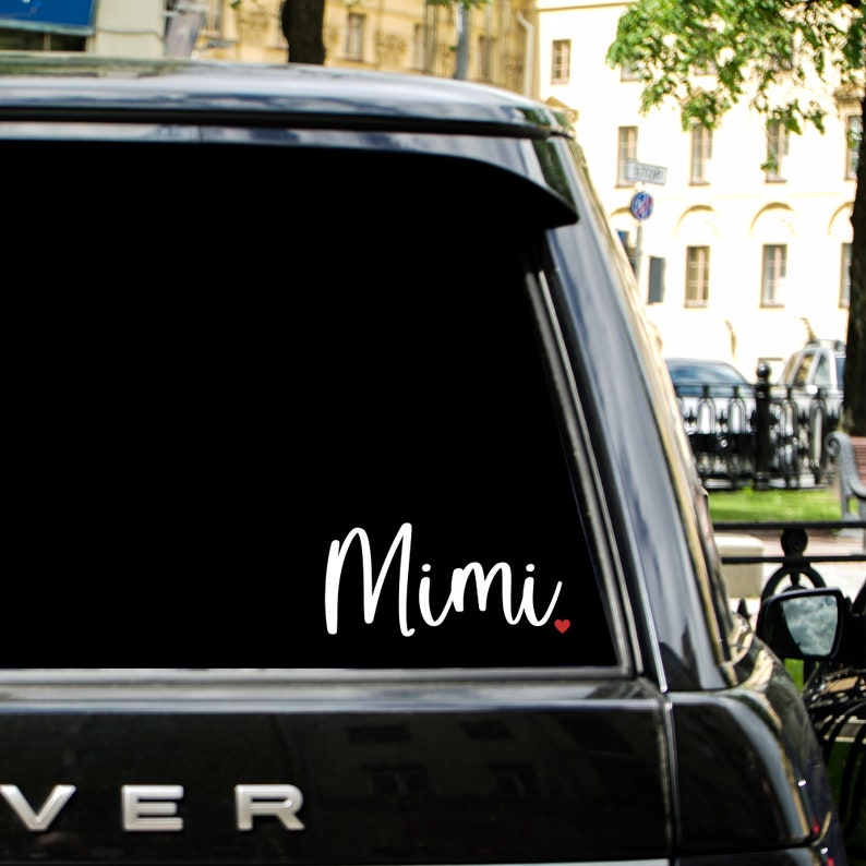 May include: White vinyl decal with the word "Mimi" in cursive script and a small red heart. The decal is on the back window of a black car.