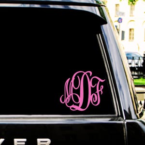 May include: A bright pink vinyl decal featuring a stylized, interlocking three-letter monogram with the letters M, D, and F in a cursive script font, applied to the dark tinted rear window of a black vehicle.