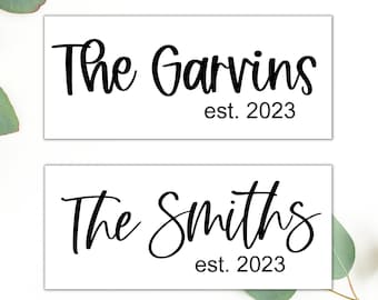 May include: Two rectangular white signs featuring black text. The top sign reads The Garvins est. 2023 in a mix of script and sans-serif fonts. The bottom sign reads The Smiths est. 2023 in the same black lettering and font styles.