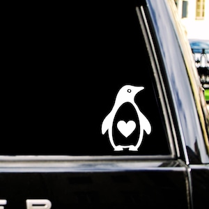 以下が含まれることがあります： 暗い車両の窓に貼られた、ペンギンのシルエットをかたどった白いビニールステッカー。ペンギンのデザインは、白い輪郭線と胸の中央にある目立つハートの形で構成されており、シンプルで明確なグラフィックディスプレイを作り出しています。
