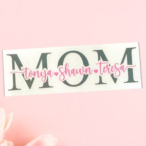 Könnte beinhalten: Ein weißes Schild mit dem Wort "MOM" in großen grauen Buchstaben. Unter den Buchstaben stehen die Namen "Tonya", "Shawn" und "Teresa" in rosa Kursivschrift mit einem kleinen rosa Herz zwischen jedem Namen.