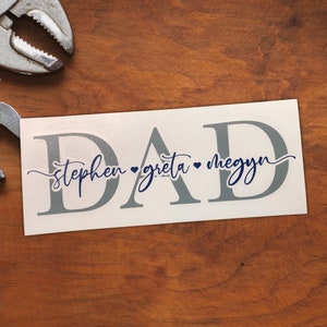 Könnte beinhalten: Ein rechteckiger weißer Aufkleber mit dem Wort DAD in großen, grauen Serif-Großbuchstaben. Überlagert in der Mitte in einer dunkelblauen Schreibschrift stehen die Namen stephen, greta und megyn, jeweils getrennt durch ein kleines Herzsymbol.