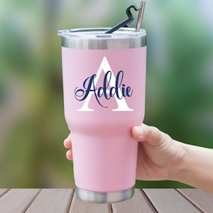 May include: A pink insulated tumbler with a silver rim and lid. The tumbler features a white letter "A" and the name "Addie" in navy blue script. A black straw and a silver straw are inserted into the lid.
