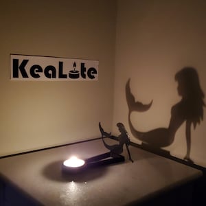 May include: A black metal mermaid candle holder with a lit tea light candle. The candle holder casts a shadow of a mermaid on the wall behind it. The text "KeaLite" is visible on the wall.